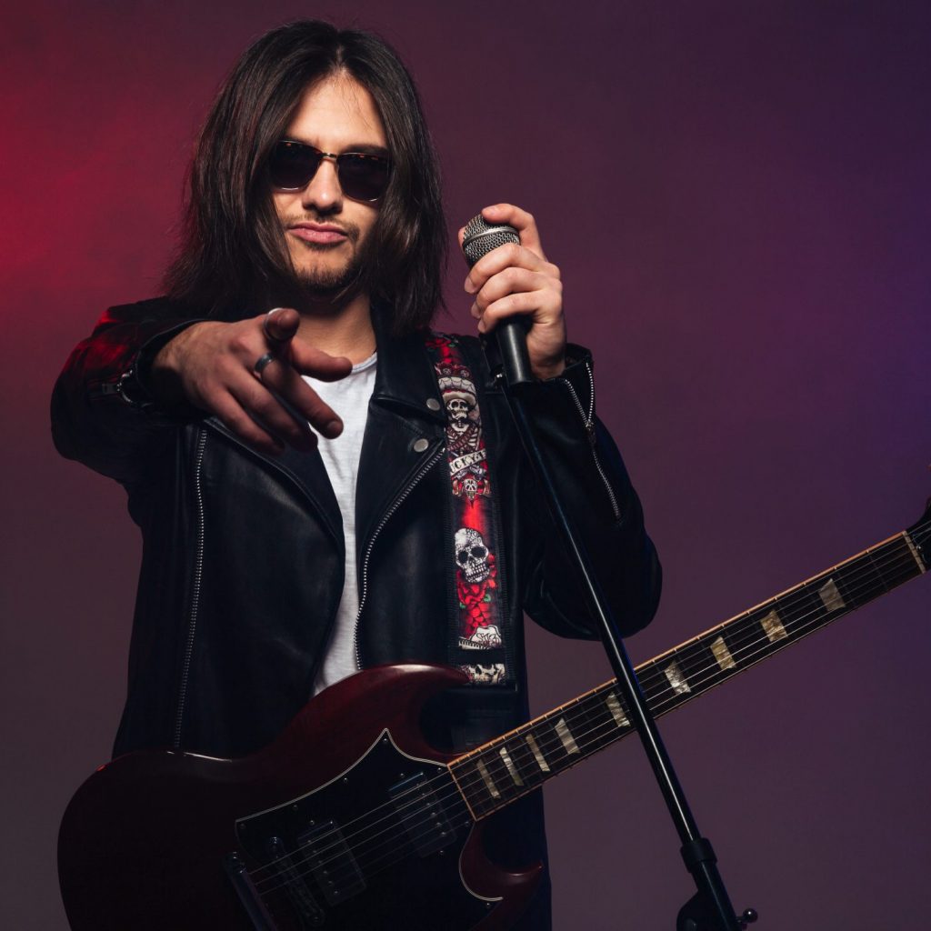 Attractive young man singer with guitar holding microphone and pointing on you over colorful smoky background